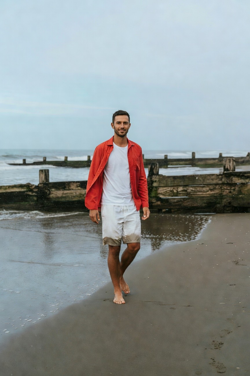 A handsome man with short dark hair walks barefoot on a sandy beach at low tide, wearing an open vibrant red jacket over a fitted white t-shirt and mid-thigh white shorts, with weathered wooden groynes and calm ocean waves in the overcast background.