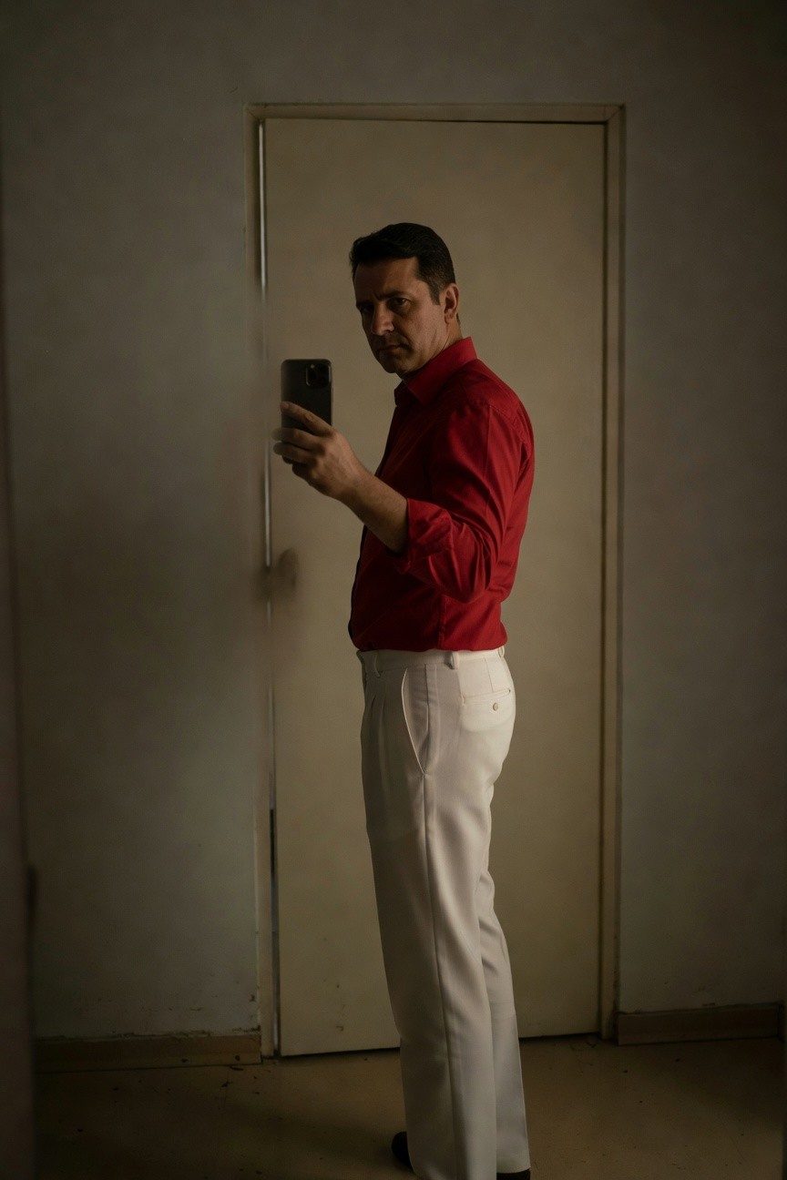 Side-profile selfie of a man in a fitted red button-down shirt with rolled sleeves and slim white trousers, standing by a plain white door in a warmly lit room, holding a phone