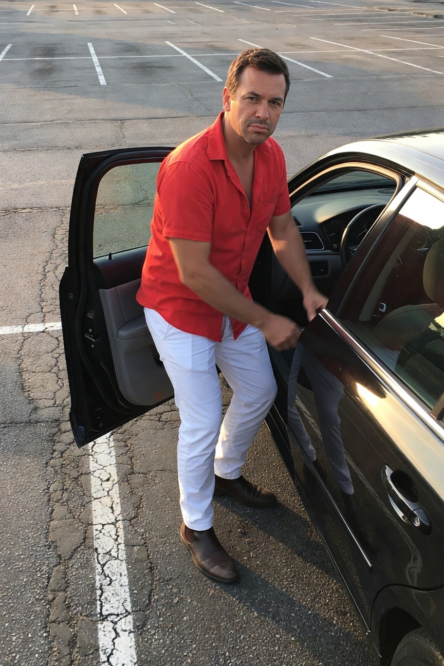 A man in a vibrant red short-sleeve button-up shirt with top unbuttoned, white slim-fit pants, and brown loafers stands confidently by the open door of a black sedan in a sunset-lit parking lot, one hand on the door frame.