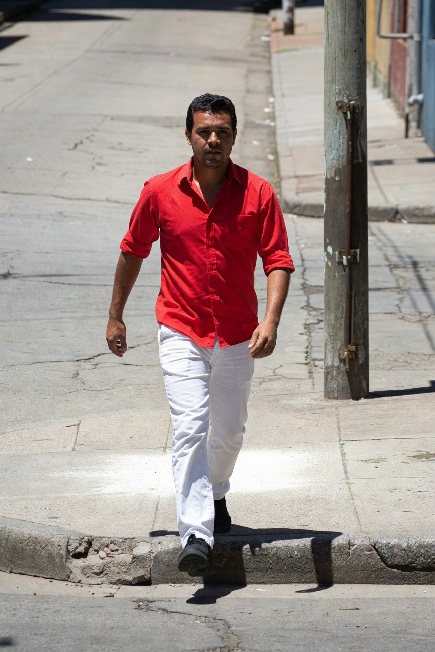 Man in vibrant red short-sleeve button-up shirt and slim white pants walking confidently on sunny urban sidewalk with pole and buildings in background