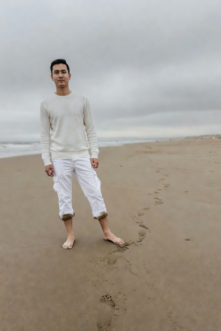 A stylish Asian man stands barefoot on a cloudy beach, dressed in a cream long-sleeve knit sweater and white cropped cargo pants with rolled cuffs, ocean waves and sandy footprints visible behind him