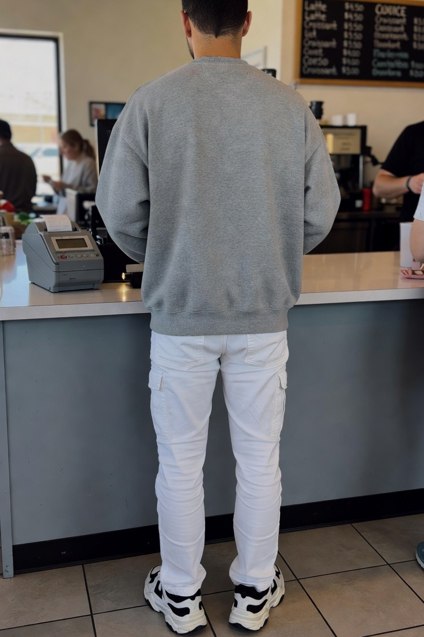 Back view of a man in a gray crewneck sweatshirt, white cargo pants, and black-and-white chunky sneakers standing at a coffee shop counter, casual daytime vibe