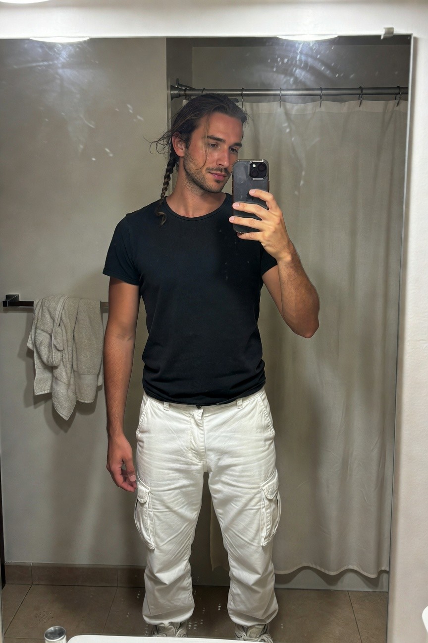 Man with long dark hair in loose braids takes a mirror selfie in a steamy bathroom, wearing a black short-sleeve t-shirt with a shoulder tear, white cargo pants, and white sneakers, standing confidently by the shower curtain