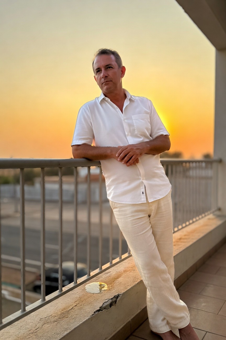 Middle-aged man in open white short-sleeve shirt and wide-leg cream linen pants stands barefoot on a balcony at sunset, leaning casually on the railing with arms crossed, cityscape and orange sky behind him