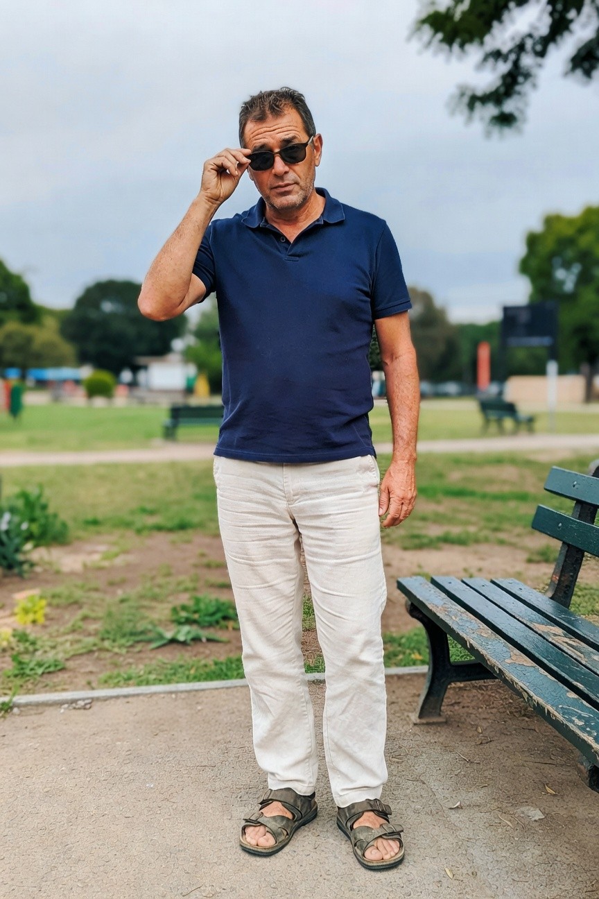 Middle-aged man in navy short-sleeve polo shirt, cream linen pants, and strappy sandals stands confidently in a grassy park under overcast skies, sunglasses perched on his head, evoking relaxed summer elegance