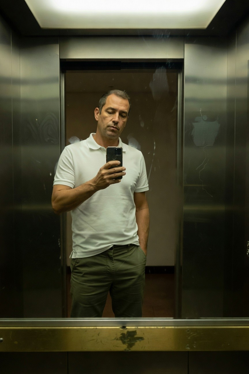 Middle-aged man in elevator mirror selfie, wearing fitted white short-sleeve polo shirt tucked into slim olive-green trousers, one hand in pocket holding phone, neutral expression, modern stainless steel elevator interior