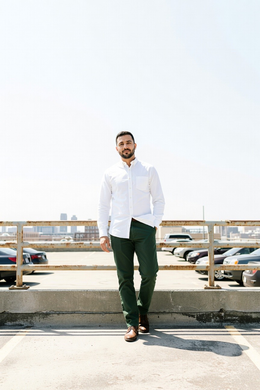 Man with dark hair and beard stands confidently on a rooftop parking structure overlooking cars and city skyline, wearing a white button-up shirt tucked into slim green trousers and brown leather shoes, sky bright blue behind him