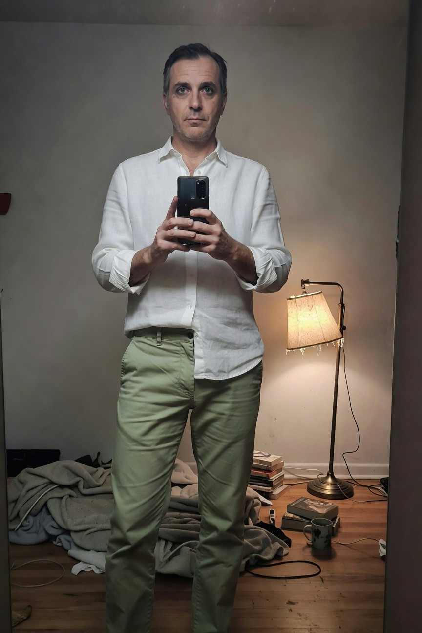 Man in white linen button-up shirt with rolled sleeves and sage green chinos stands casually in a dimly lit room, holding phone for selfie amid scattered clothes and books on wooden floor