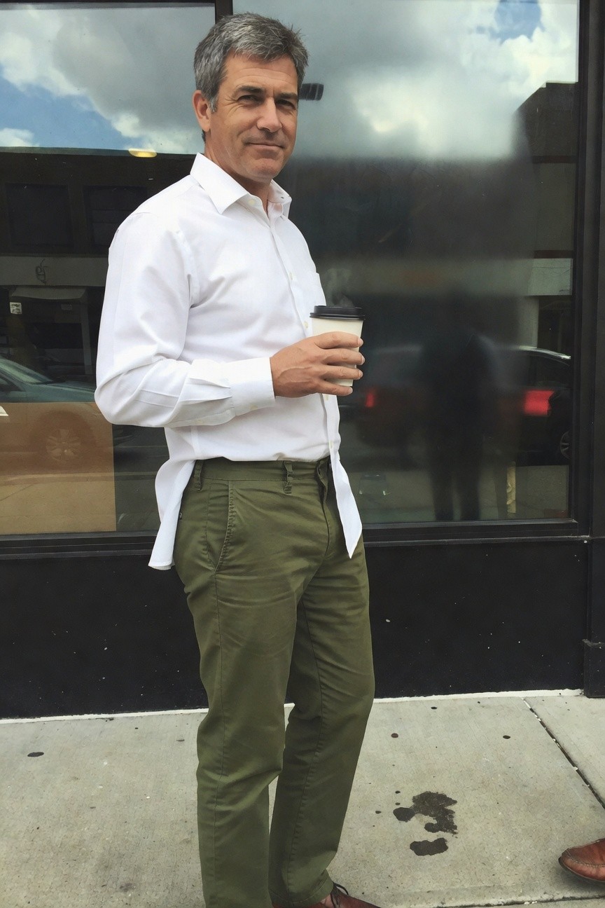 Middle-aged man in slim olive green chinos, white long-sleeve button-down shirt half-tucked asymmetrically, brown loafers, holding a to-go coffee cup while standing casually outside a cafe window on a cloudy day