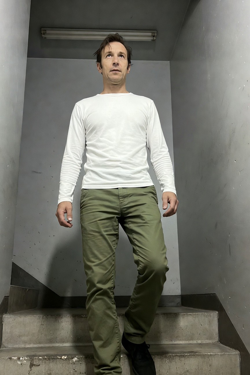 Man in white long-sleeve shirt and slim olive green chinos stands mid-stride on concrete stairs in a dimly lit stairwell, black sneakers on steps, hands relaxed at sides