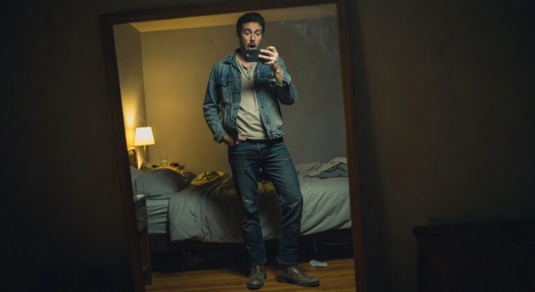 Man taking a mirror selfie in a light wash denim jacket over a cream henley shirt, dark blue straight-leg jeans, and brown leather boots, standing confidently in a bedroom setting