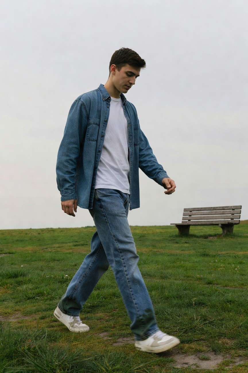 Young man walking in casual Western-inspired outfit: open blue denim shirt over white t-shirt, blue jeans, white sneakers on grassy path