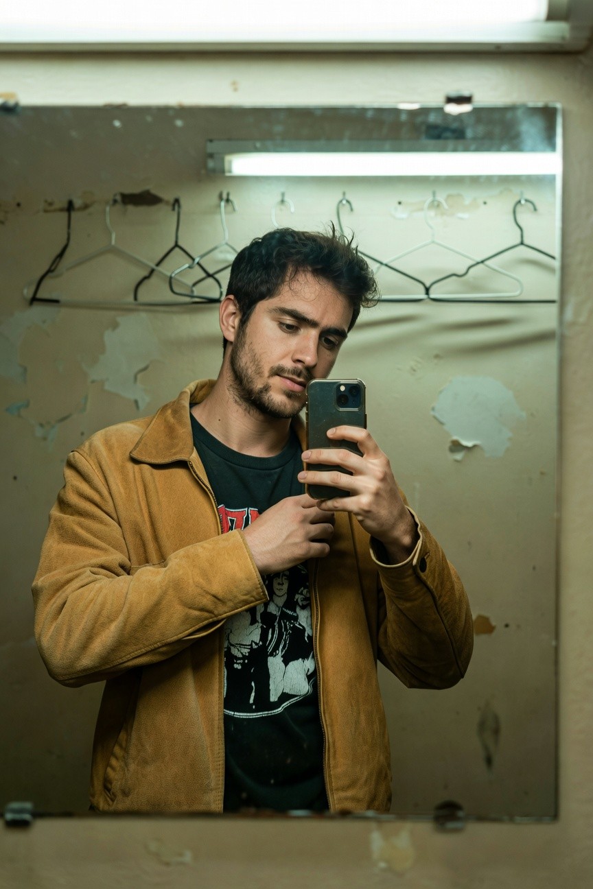 Man taking mirror selfie in worn bathroom, wearing open tan corduroy jacket over black graphic t-shirt with white face print, dark hair and beard, casual rugged style