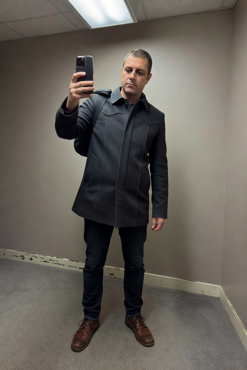 Man in dark gray wool Western-style coat with shoulder yoke, slim dark jeans, and brown leather boots, posing in a beige fitting room