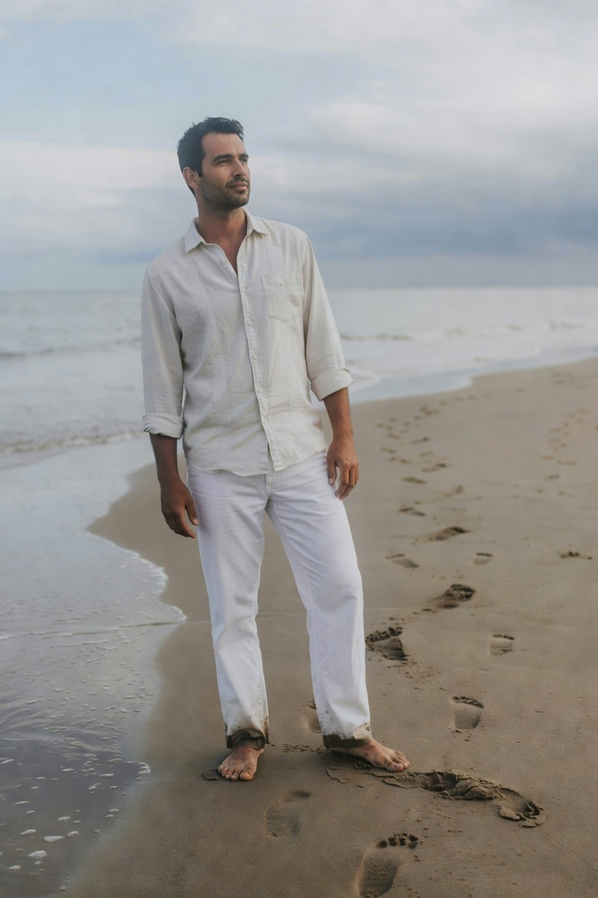 Man standing on sandy beach in rolled-sleeve white linen shirt, matching wide-leg pants, barefoot, with ocean waves nearby