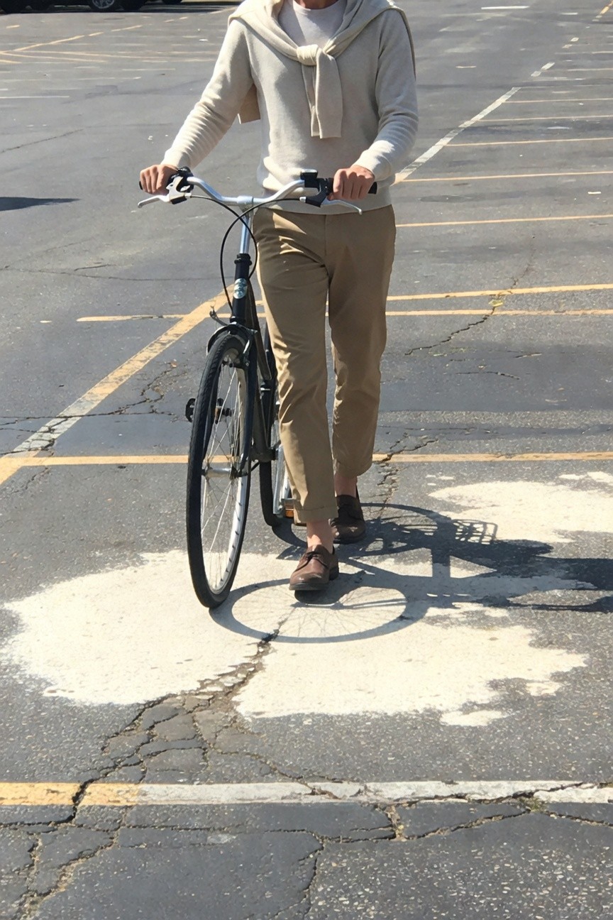 Man biking in empty parking lot wearing cream sweater with draped scarf, light khaki tailored ankle pants, brown loafers, black upright bicycle, sunny day with shadows on asphalt