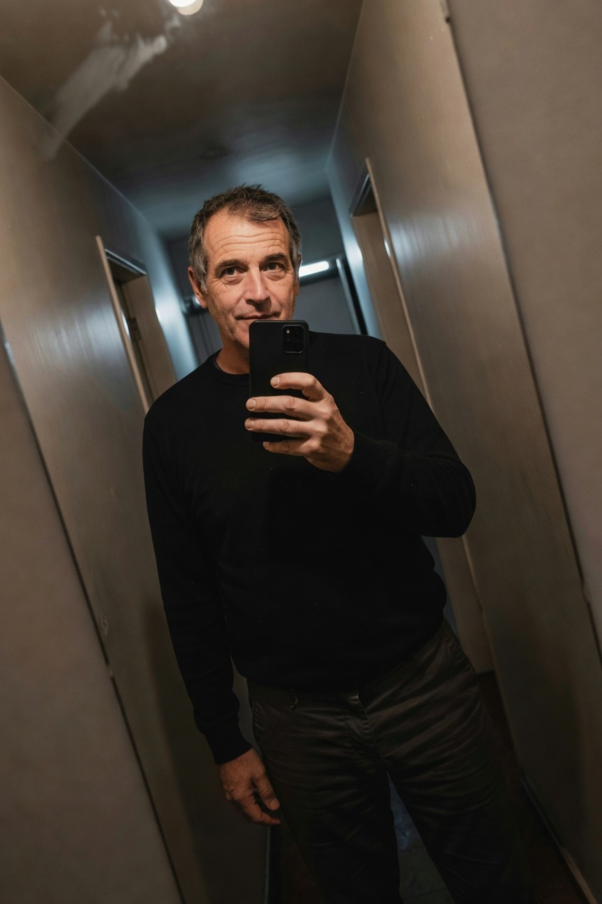 Man in black fine-knit sweater and gray straight-leg trousers poses confidently in a mirrored hallway, holding phone for selfie, warm lighting highlighting casual sophistication