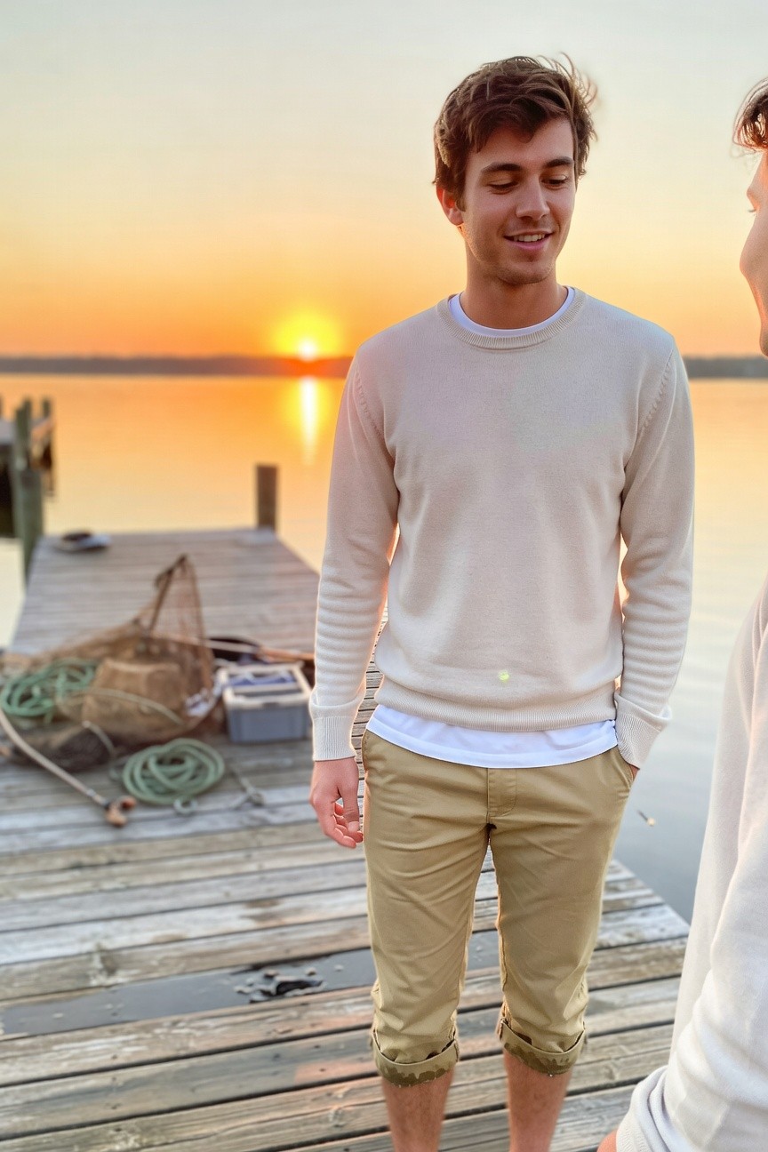 A young man stands confidently on a wooden dock at sunset, wearing a light beige knit sweater over a white t-shirt, khaki chinos cropped at the ankles, barefoot, with calm water and fishing nets in the background, evoking relaxed coastal style.