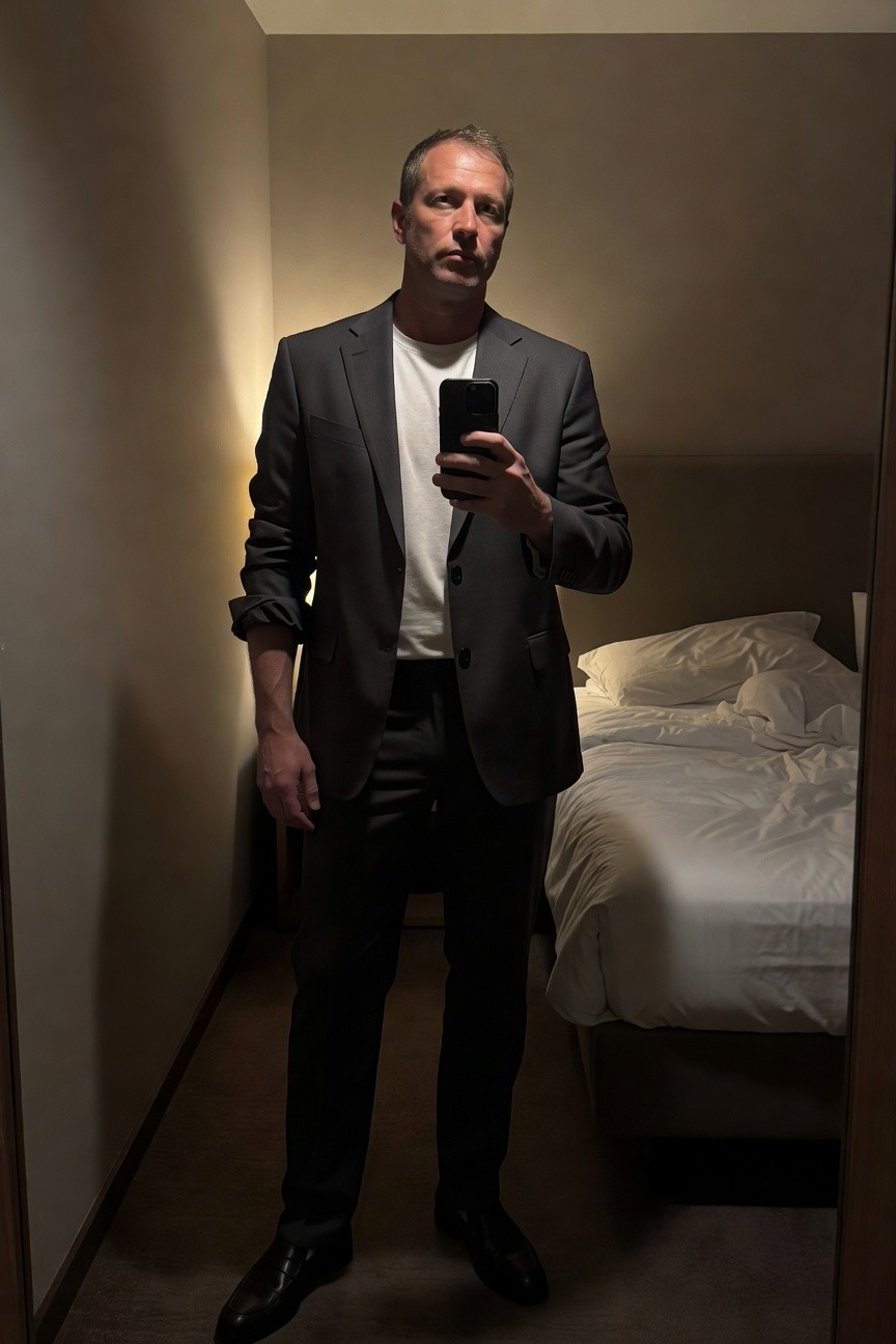 Man in slim-fit charcoal gray suit jacket and trousers over white crewneck t-shirt, black dress shoes, standing in dimly lit hotel room with bed in background, selfie pose holding phone