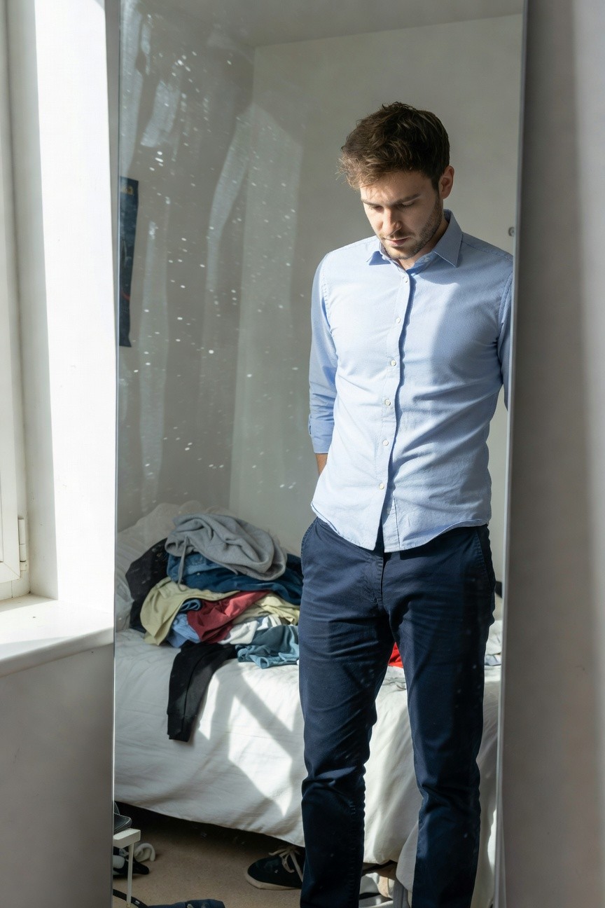 Man standing in front of mirror wearing light blue long-sleeve button-up shirt tucked into slim navy trousers and dark sneakers, casual bedroom background with laundry on bed