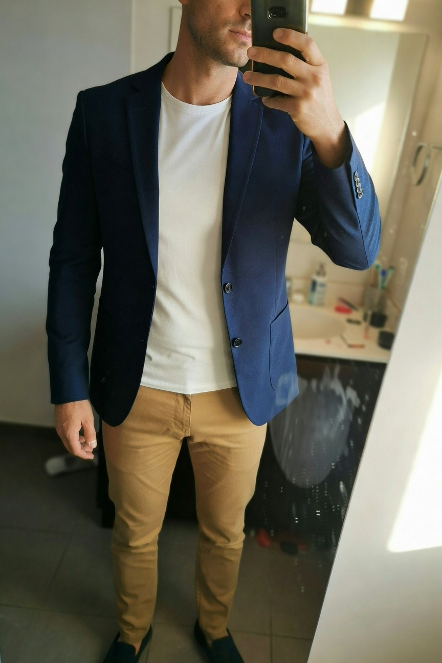 Man in slim navy blazer over white crewneck t-shirt, beige slim-fit chinos, and black slip-on shoes, taking a mirror selfie in a bathroom with sink visible