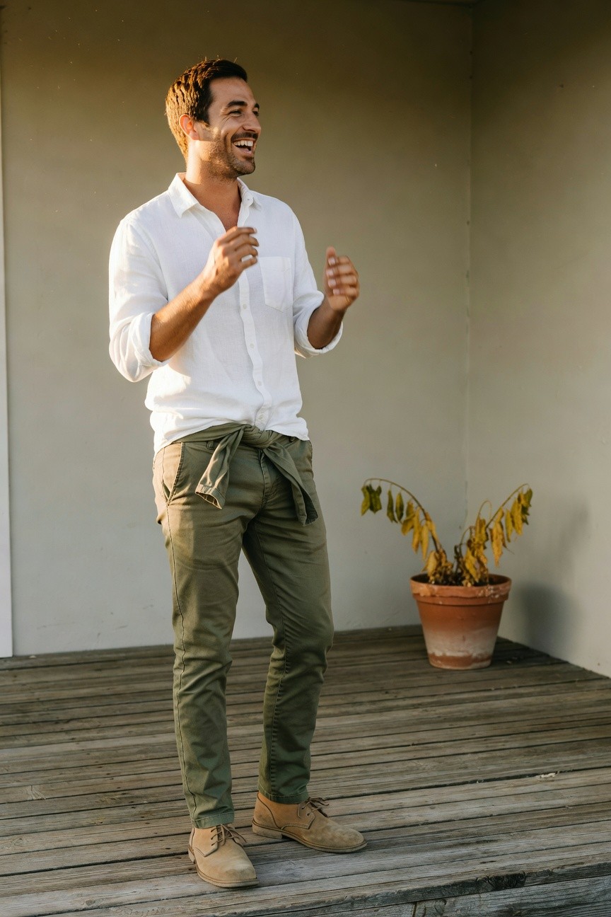 Man in white long-sleeve button-up shirt gesturing with hands, olive green straight-leg pants tied at waist with fabric belt, tan suede chukka boots, standing on wooden deck beside potted plant