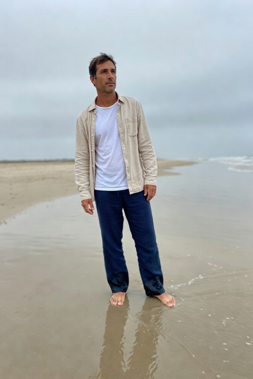 Man in open light beige button-up shirt over white t-shirt and relaxed navy pants stands barefoot on wet beach sand under overcast sky, evoking serene summer casualness