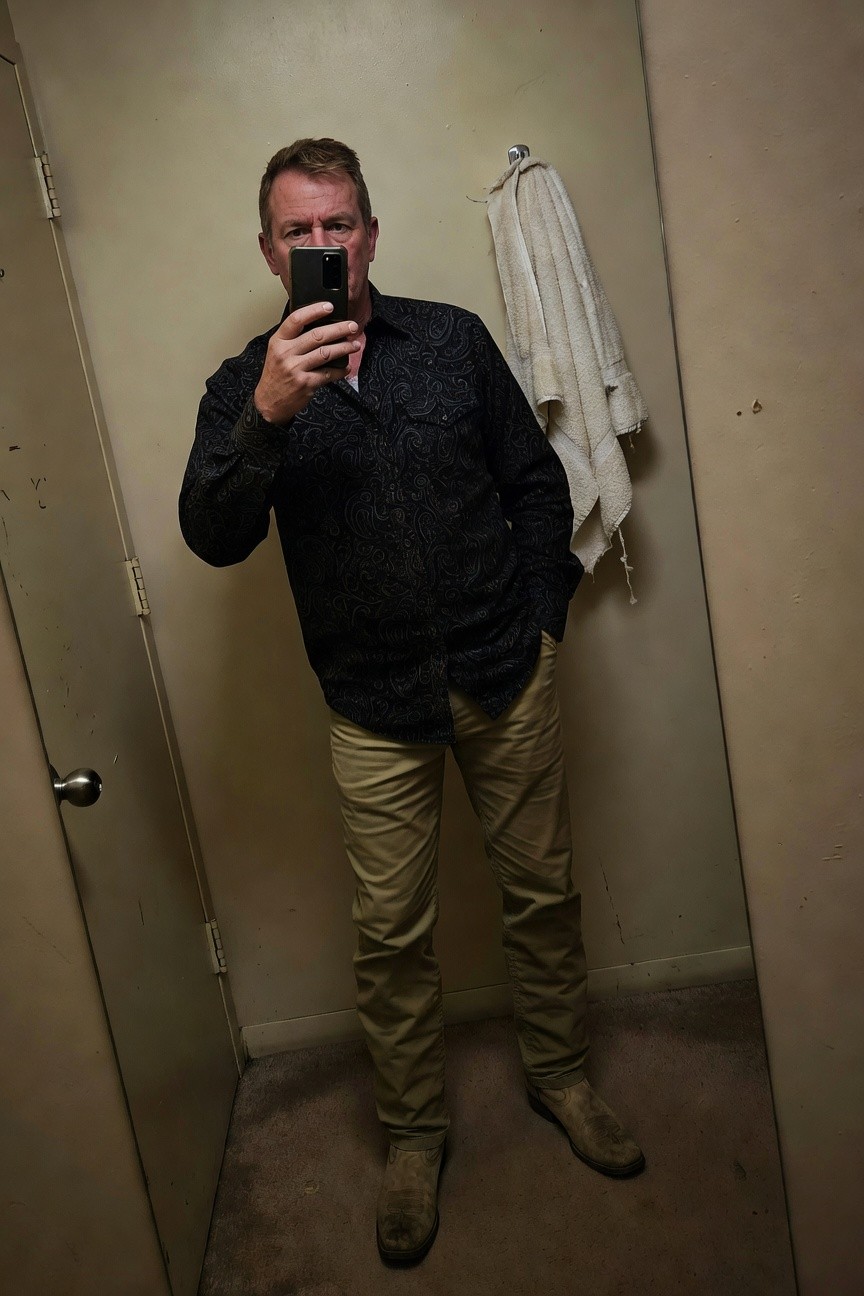 Man in bathroom mirror selfie wearing dark patterned button-up shirt tucked into khaki chinos and light suede desert boots, white towel hanging on door hook behind him