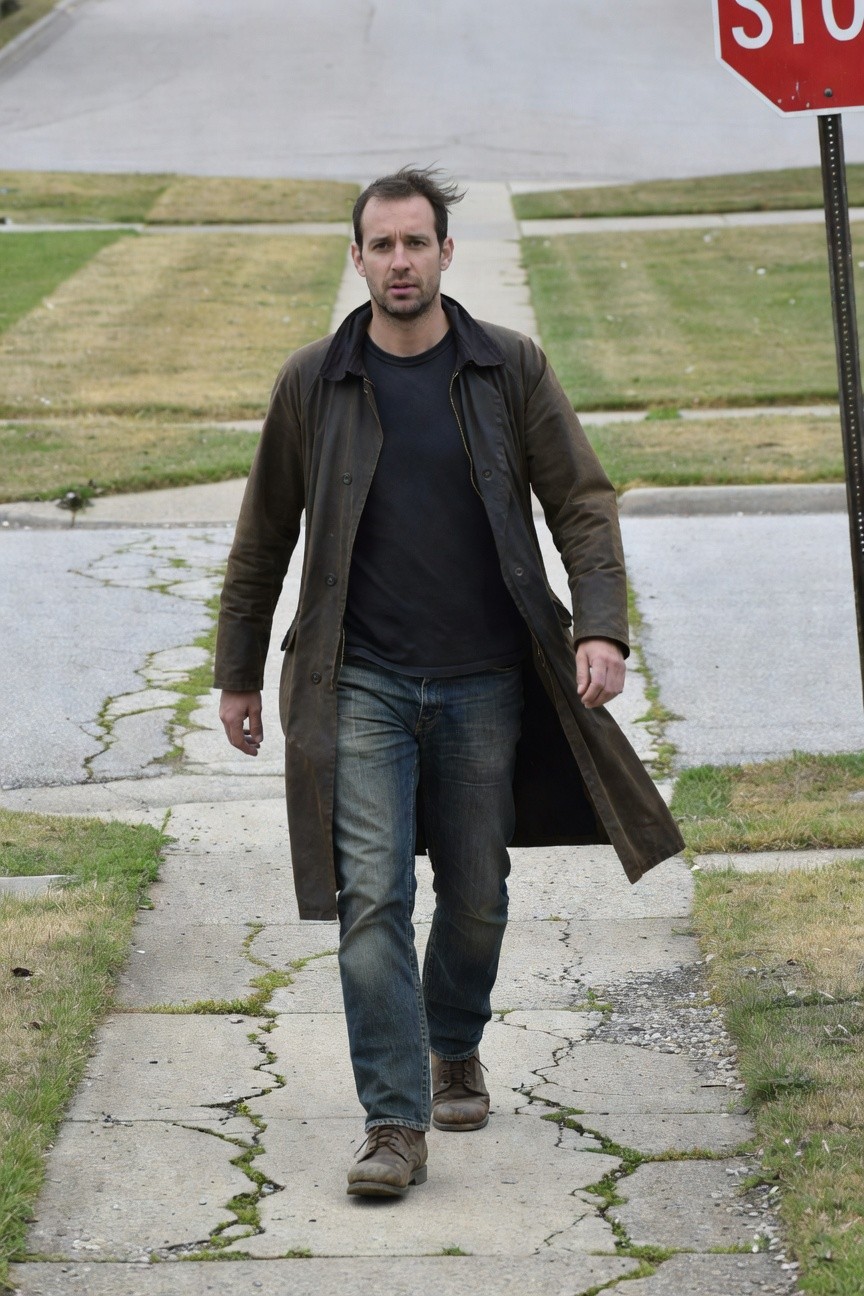 Man in long brown duster coat over black t-shirt and blue jeans with brown boots walking on sidewalk