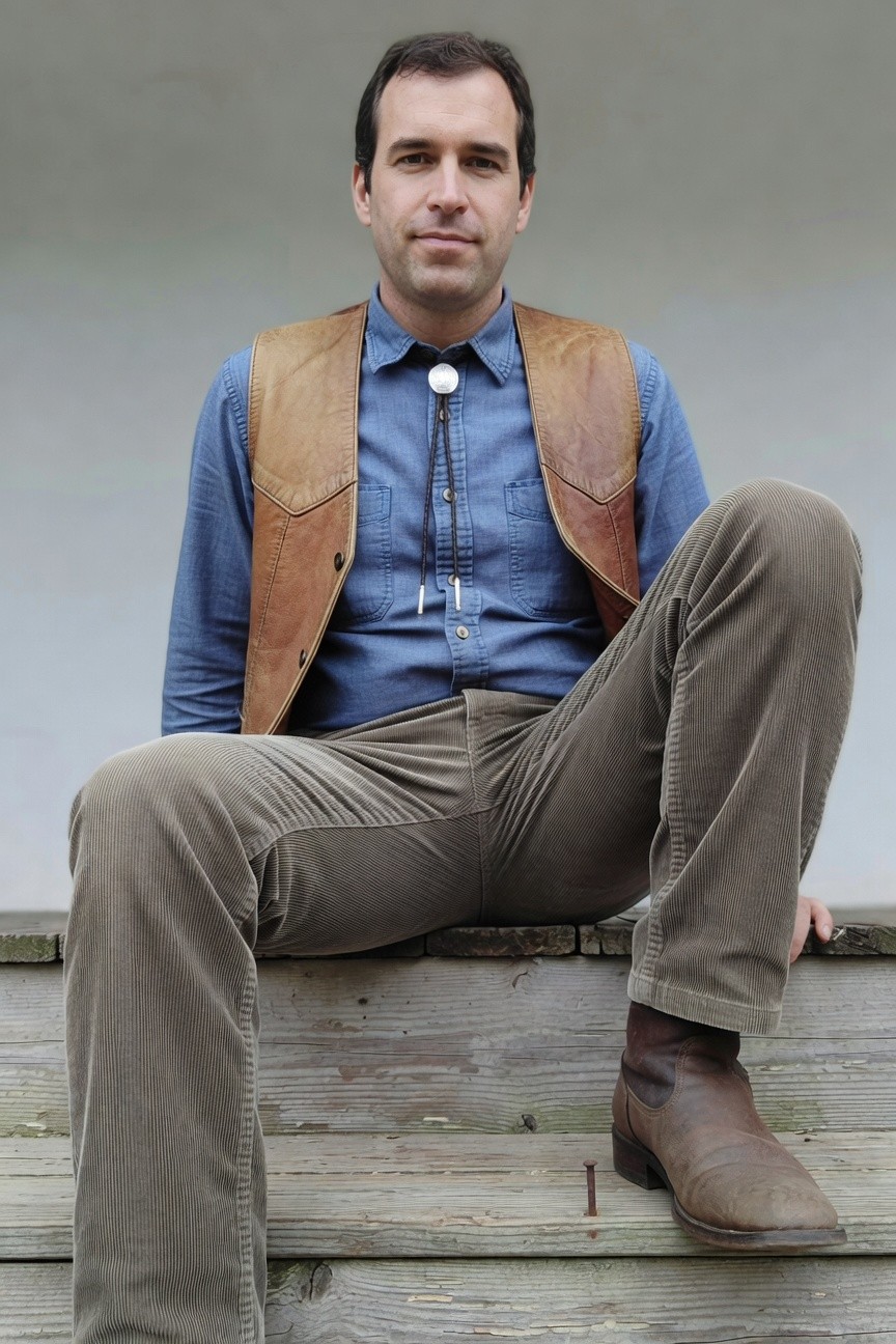 Man seated on wooden steps in a blue denim shirt layered under a brown leather vest, navy bolo tie, gray corduroy pants, and tan cowboy boots, gray background