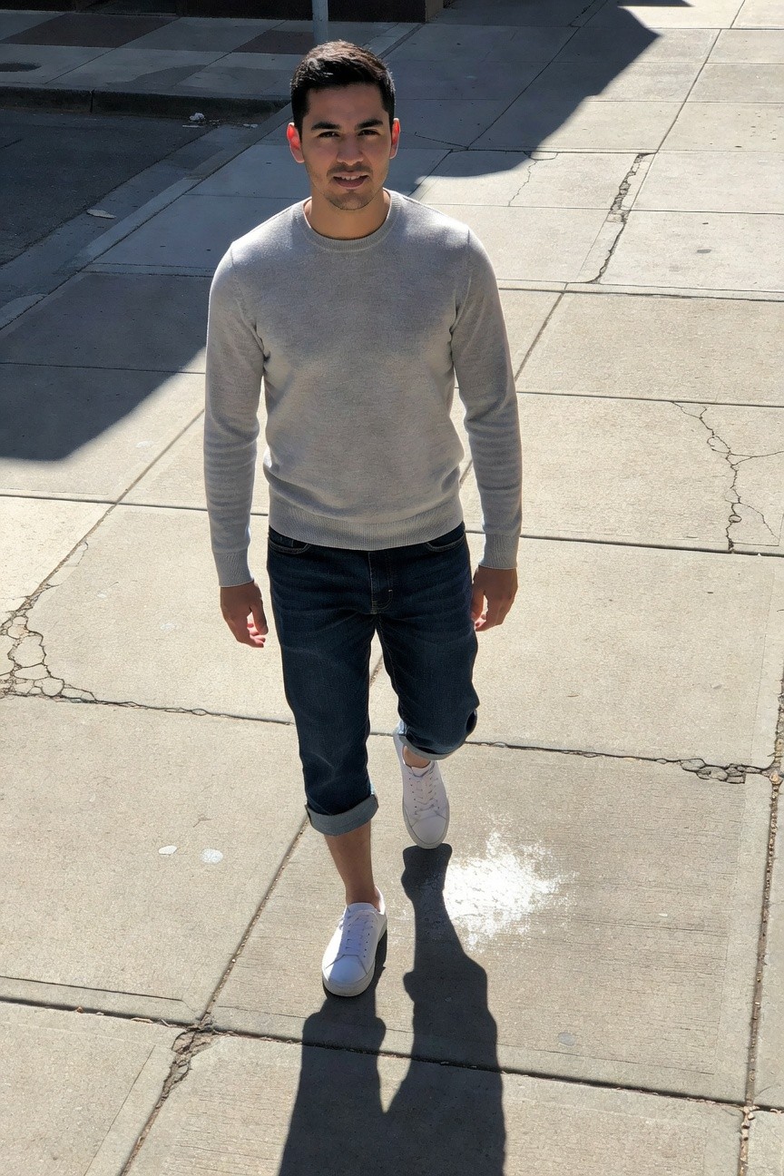 Man in light gray long-sleeve crewneck sweater, dark rolled-up jeans to mid-calf, white low-top sneakers, walking confidently on sunny cracked sidewalk with shadow trailing behind