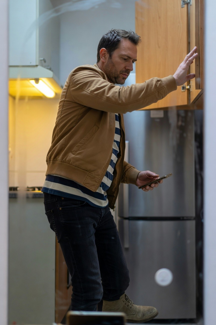 Man in open tan suede jacket over striped shirt, slim dark jeans, and tan boots stands in modern kitchen reaching into wooden cabinet while holding phone, warm yellow light glowing behind