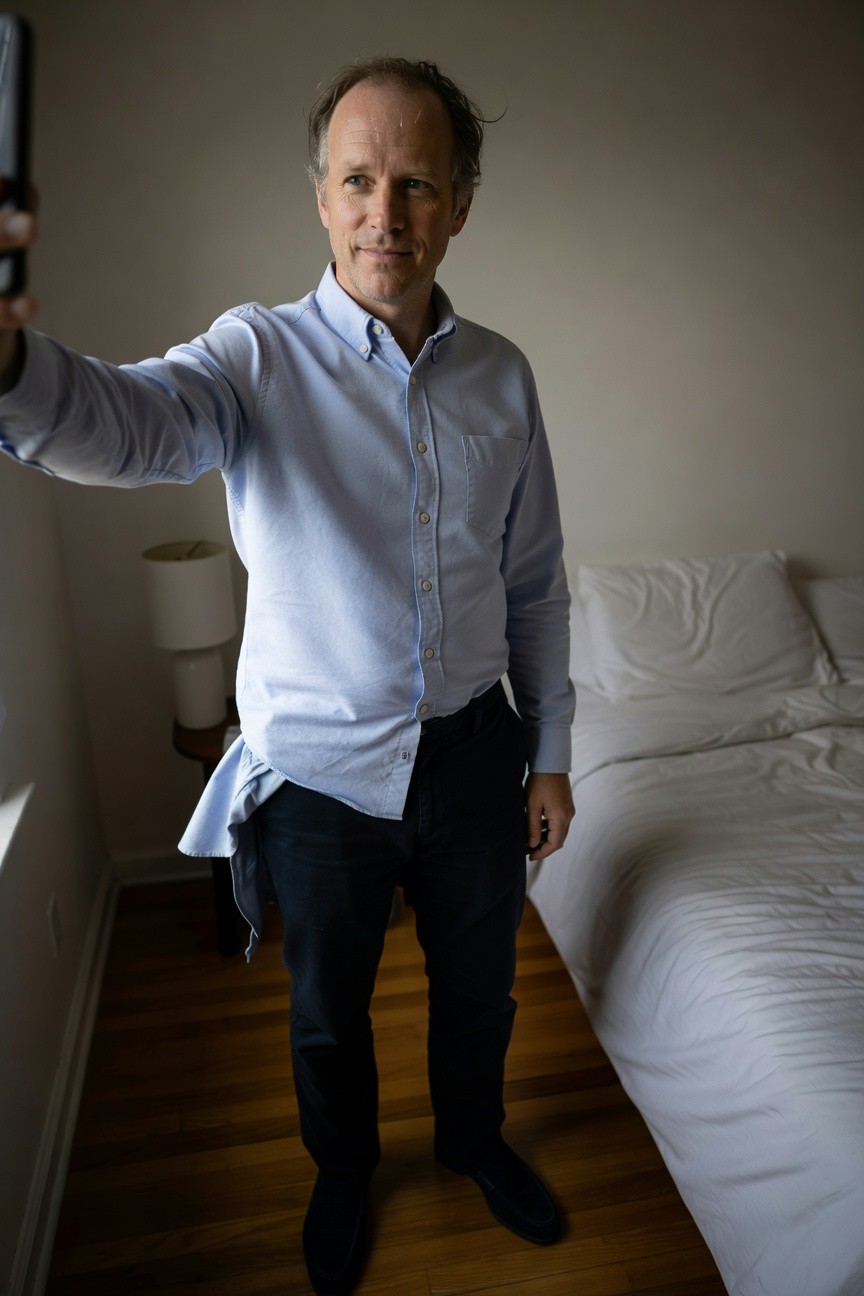 Middle-aged man in light blue untucked button-down shirt, slim navy trousers, and dark loafers, taking a selfie in a cozy bedroom with white bed and lamp in background