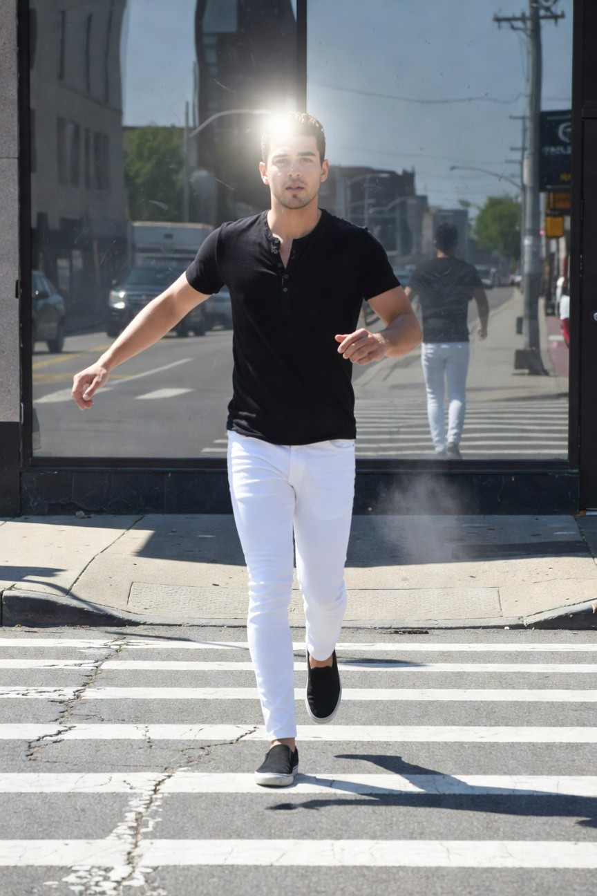Man in black short-sleeve polo shirt with rolled sleeves and slim white jeans strides confidently across an urban crosswalk, white sneakers on feet, bright sunlight flaring behind him against a glass storefront
