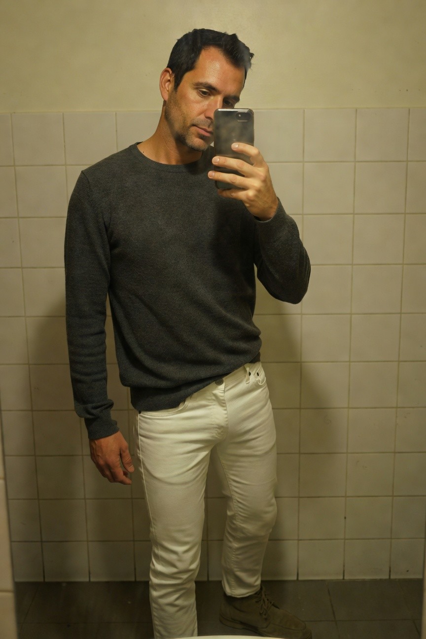 Man in charcoal gray crewneck sweater and slim white jeans stands casually with hands in pockets in front of bathroom mirror, wearing light tan suede shoes, selfie-style photo