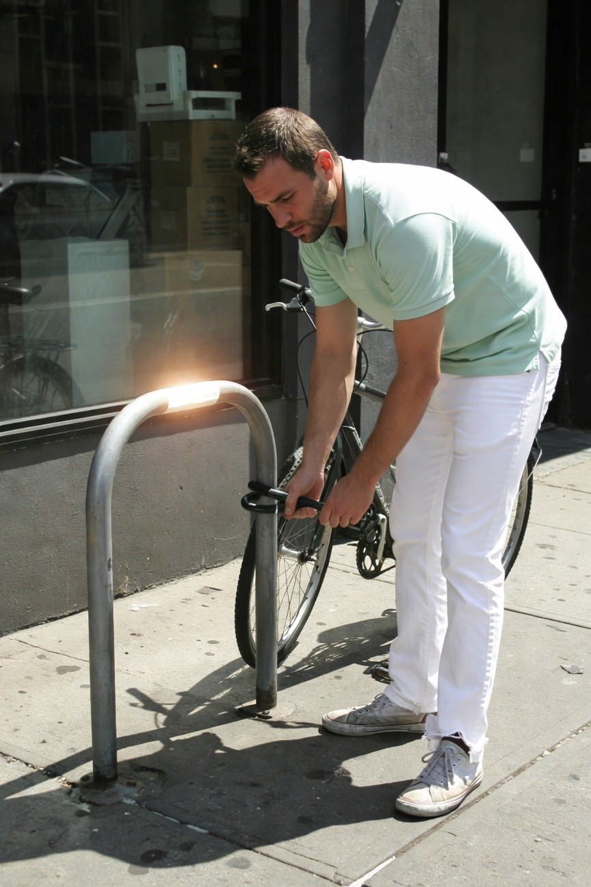 Man in pale mint green short-sleeve polo shirt and slim white jeans bends to lock black road bike to curved metal rack on sunny urban sidewalk, white sneakers on littered pavement, glass building backdrop
