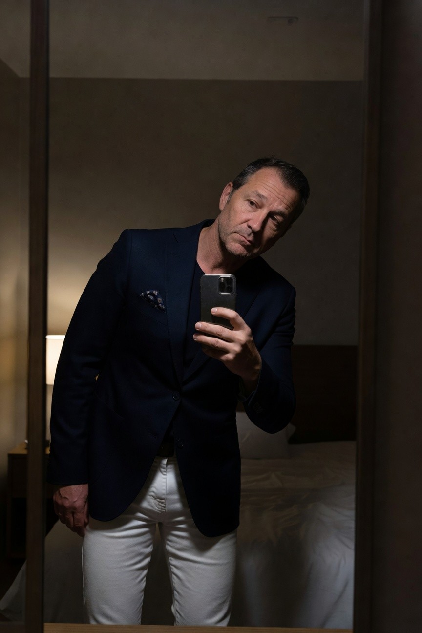 Man in navy blazer with pink pocket square over black turtleneck, crisp white jeans, holding phone in hotel room mirror selfie