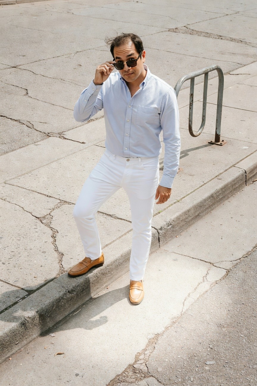 Man in light blue long-sleeve button-up shirt, slim white jeans, and brown leather loafers poses confidently on sunlit urban street near metal bike rack, sunglasses in hand