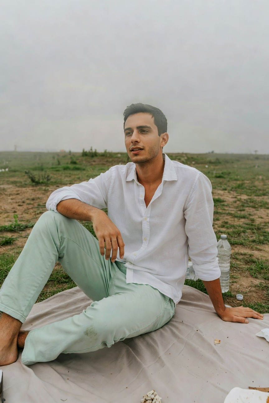 A man sits barefoot on a beige picnic blanket in grassy field under cloudy sky, wearing slim light mint green pants and a loose white linen long-sleeve shirt unbuttoned at collar, one knee up gazing sideways pensively.