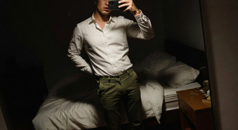 Man taking mirror selfie in bedroom wearing white long-sleeve button-up shirt tucked into olive green chinos, hands in pockets, bed and lamp in background