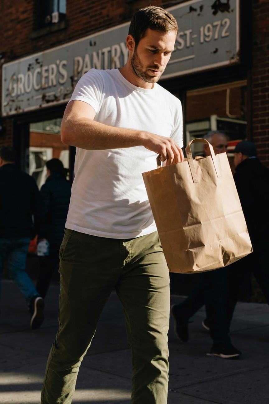 A fit man with short dark hair stands on a bustling sidewalk outside Grocer's Pantry, wearing a fitted white cotton t-shirt tucked loosely into slim olive green chinos, holding a full brown paper grocery bag; casual street scene with pedestrians and store signage in background.