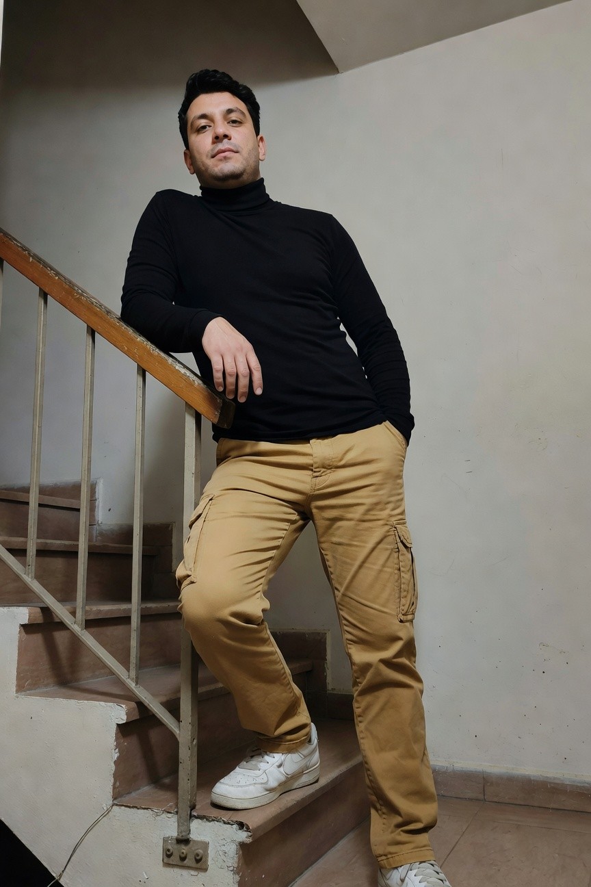 Man in black long-sleeve turtleneck sweater leaning on wooden staircase railing, wearing beige khaki cargo pants with multiple pockets and white Nike Air Force 1 sneakers, casual indoor pose