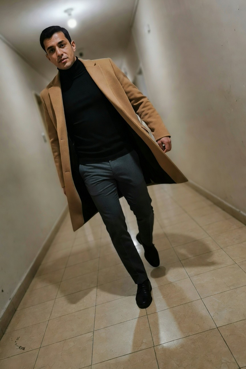 Man striding down an indoor hallway in an open camel-colored overcoat over a black turtleneck sweater, slim gray trousers, and black leather dress shoes