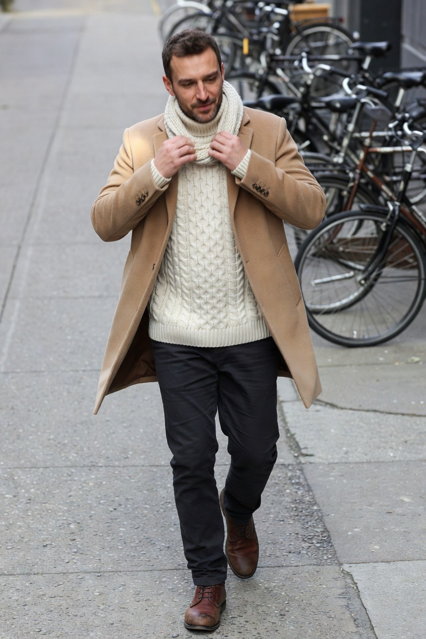 Man walking on sidewalk wearing open camel wool overcoat over white cable knit turtleneck sweater, black slim trousers, and brown leather boots, adjusting sweater collar with row of bicycles in blurred background
