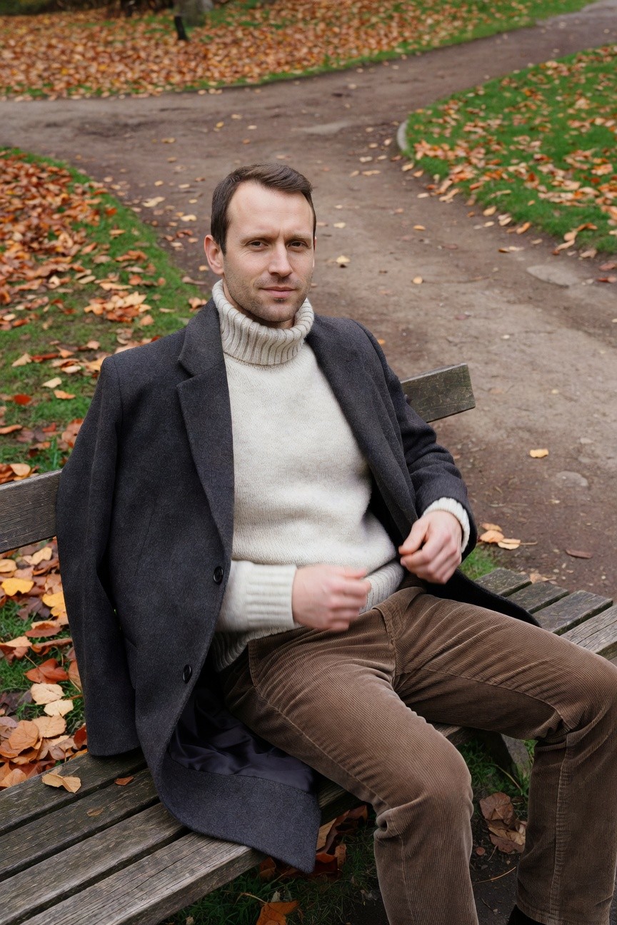 Man seated on wooden bench in cream cable-knit turtleneck sweater layered under open charcoal gray wool overcoat with brown corduroy trousers and black shoes, hands adjusting sweater hem against blurred park path backdrop