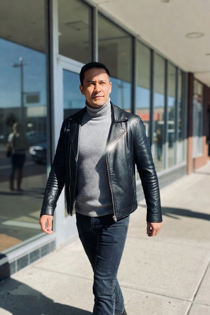 Man in gray ribbed turtleneck sweater layered under black zip-up leather jacket with stitching details, paired with slim dark jeans and black shoes, walking on urban sidewalk