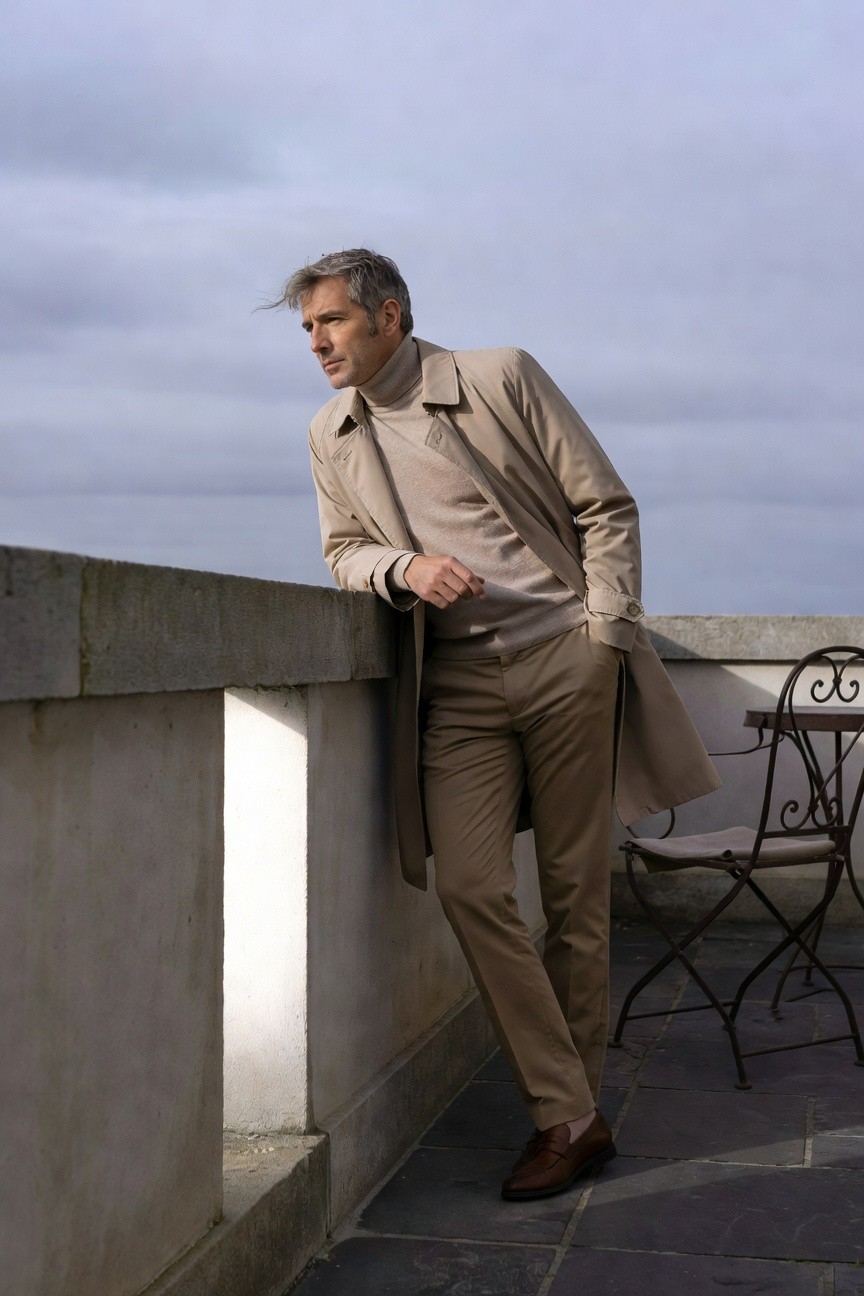 A man with silver hair wearing a cream turtleneck sweater under a beige trench coat and matching trousers, leaning against a stone wall with brown loafers, on a terrace.