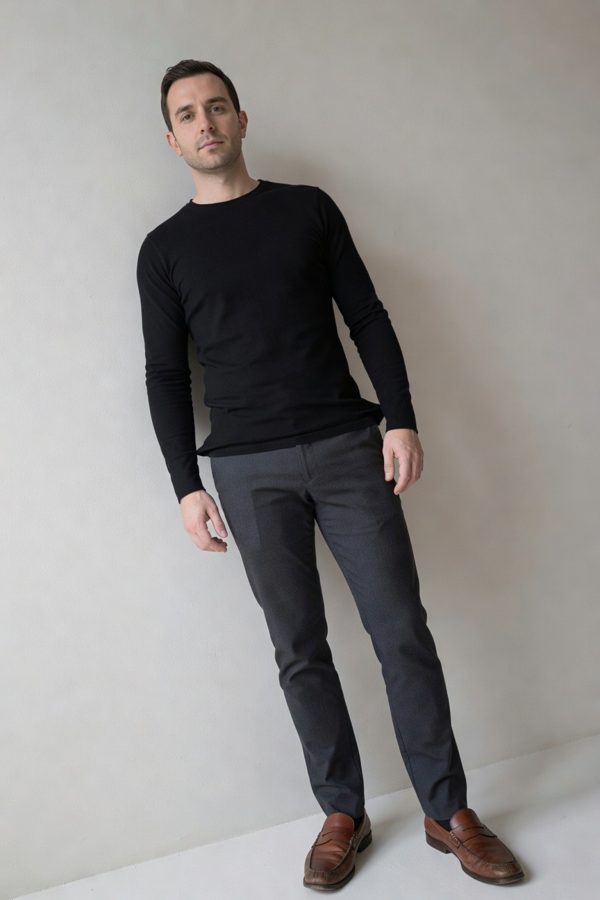 Man in black long-sleeve crewneck sweater, charcoal grey slim trousers, and brown leather loafers, leaning casually against a plain wall for a smart casual look