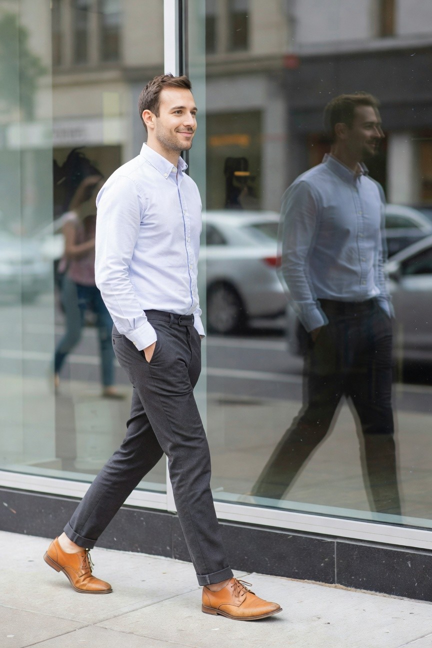 Man in light blue button-up shirt, slim charcoal grey pants cuffed at ankles, and brown leather lace-up shoes, walking past a glass storefront with his reflection visible