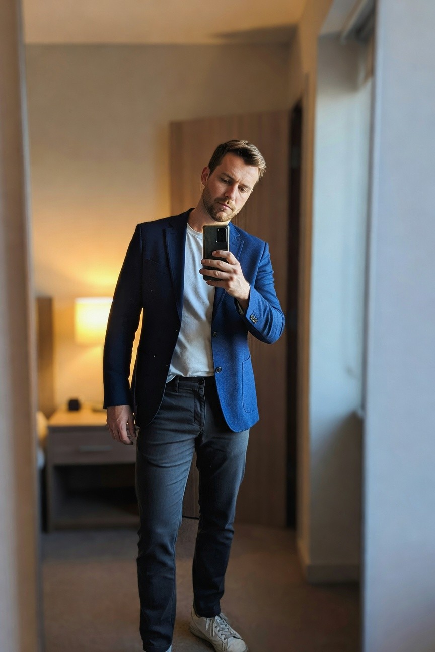 Man in navy blue blazer over white t-shirt, charcoal grey slim pants, and white sneakers, posing in front of mirror for smart casual look